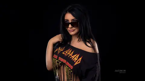 MissyJolie poses with long black hair and dark sunglasses, wearing a black graphic t-shirt against a black background.