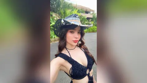 Young woman in a black crochet bikini and wide-brimmed straw hat posing outdoors in a tropical summer setting.