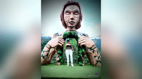 Woman in white pants standing before a massive stone goddess statue in a lush green landscape.