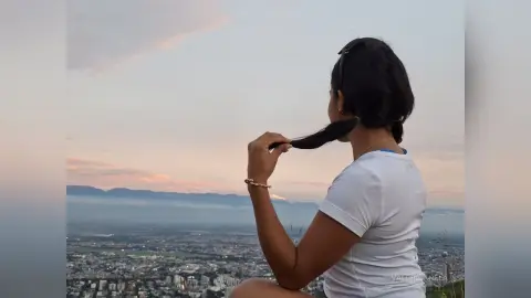 Woman in a white t-shirt looking at a scenic city view during a peaceful sunset from a mountain lookout.