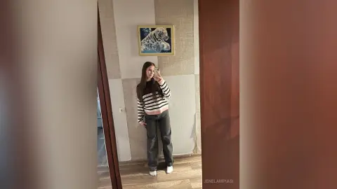 Young woman in a striped long-sleeve shirt and dark jeans taking a mirror selfie in a modern minimalist room.