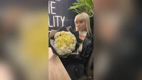 Blonde woman in a black leather jacket holding a large bouquet of white and yellow flowers in a modern cafe.