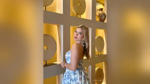 Blonde woman in a blue floral dress posing in a warm, minimalist interior with decorative wall niches.