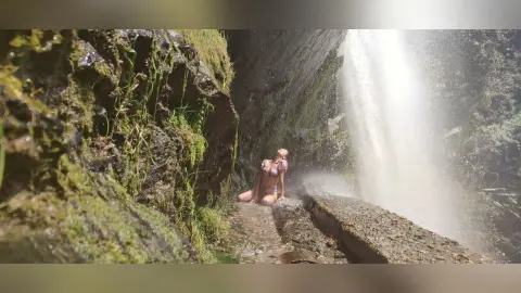 LaurentBelluci poses naked or in light pink lingerie under a waterfall, sitting on a wet rock surrounded by mossy stone walls.