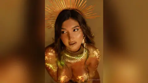 Ethereal goddess in gold celestial headpiece and jewelry with shimmering body glitter in warm lighting.