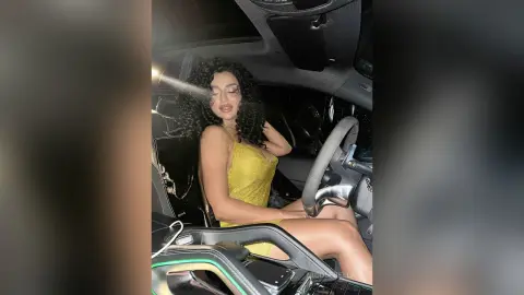 NicoleBella poses in the driver's seat of a car wearing a tight yellow dress and showing her bare legs.