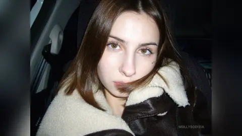 Young woman with long brown hair wearing a cozy shearling jacket sitting in a car at night.