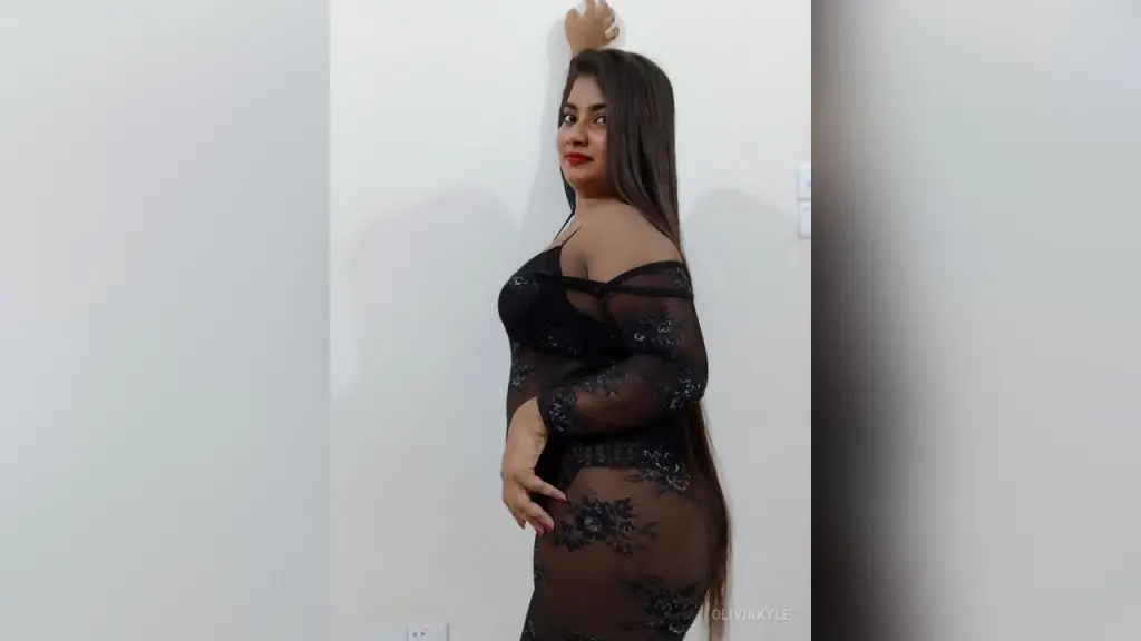 OliviaKyle — Elegant woman in a black lace sheer dress posing against a white wall in a minimalist indoor setting.