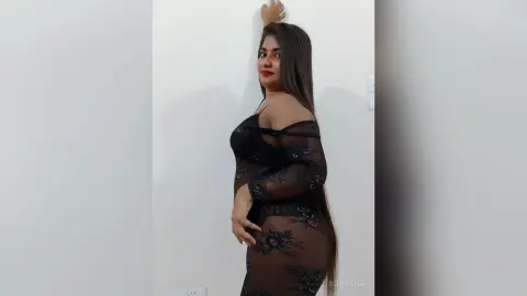 Elegant woman in a black lace sheer dress posing against a white wall in a minimalist indoor setting.