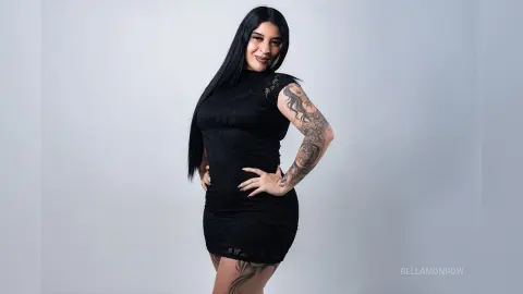 BellaMonrow poses in a tight black lace dress, showing off her long black hair and detailed arm tattoos against a plain white background.