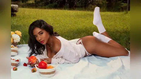 Beautiful woman in white loungewear relaxing on a picnic blanket in a sunny grassy field with fresh strawberries.