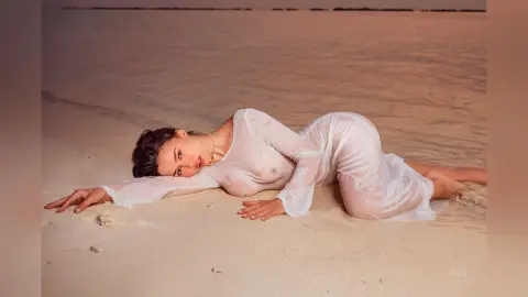 Elie lies naked on a sandy beach, wearing a sheer white long-sleeve top that shows her nipples through the wet fabric.