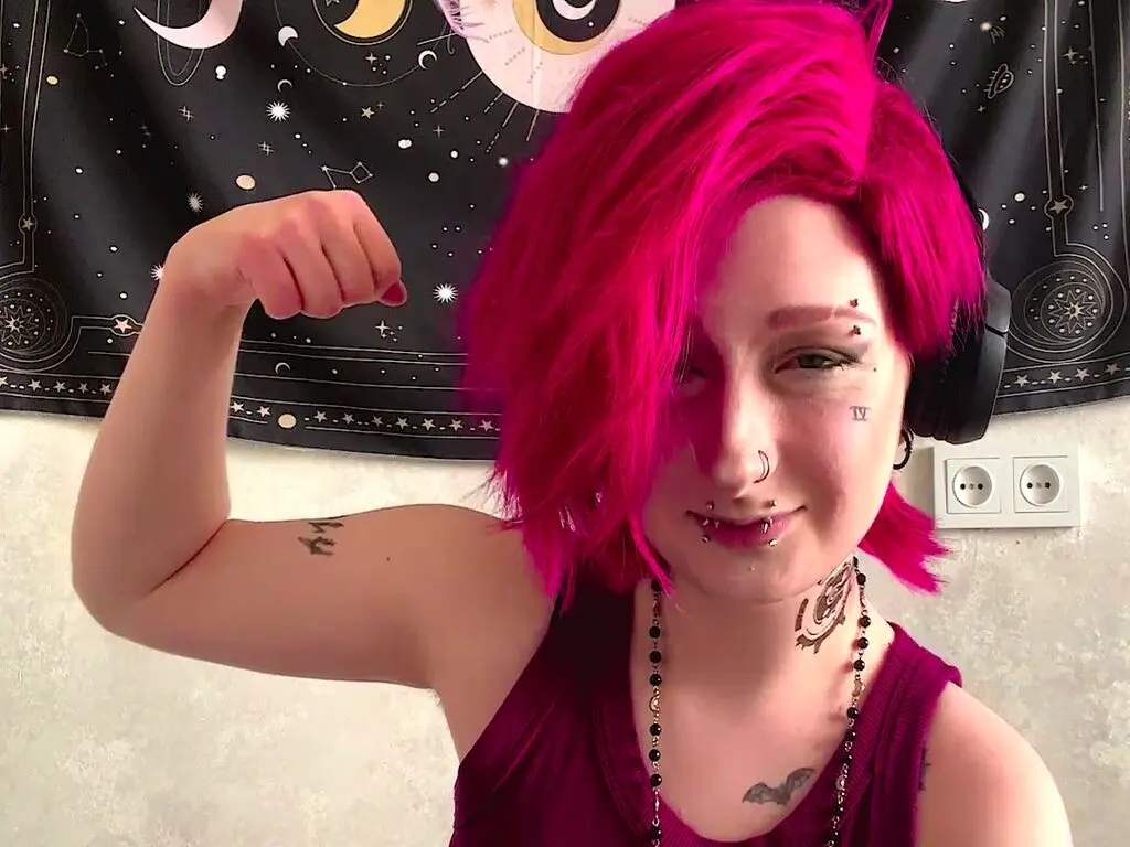 Woman with bright pink hair and tattoos flexing her arm in a casual tank top against a celestial tapestry.