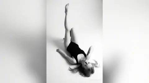 SophiePeachy lies on a white floor wearing a black bodysuit with her legs stretched up in the air.
