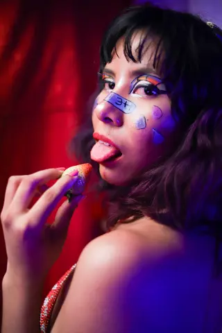 Young woman with artistic face paint eating a strawberry in a moody, red-lit setting.