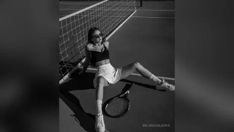 MilanaVolkova poses on a tennis court wearing a black sports bra and white skirt, leaning against the net with a racket at her feet.