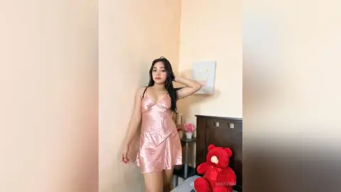Young woman in a pink satin slip dress posing in a cozy bedroom with a red teddy bear.