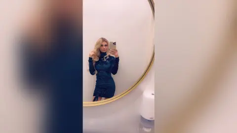 Blonde woman in a glamorous sparkling navy blue long-sleeve dress taking a selfie in a modern bathroom mirror.