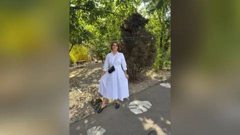 Woman in a white midi dress walking through a sunny garden setting with lush green trees.