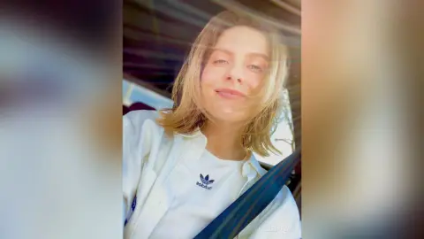 Smiling blonde woman in a white shirt sitting in a car during a bright, sunny day.