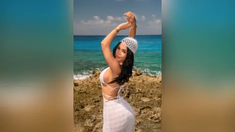 Elegant woman in a white lace crochet dress posing on rocky cliffs by the turquoise ocean under a sunny sky.