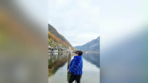 Young woman in a blue faux fur coat standing by a scenic mountain lake under a cloudy sky.