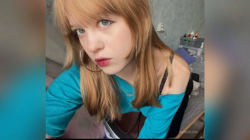 Blonde woman with bangs and silver hoop earrings wearing a teal top in a casual indoor setting.