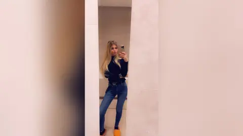 Blonde woman in a black turtleneck and blue jeans taking a mirror selfie in a minimalist modern hallway.