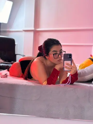 Young woman in red velvet outfit and glasses taking a mirror selfie on a bed with pink moody lighting.