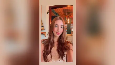 Smiling young woman with long wavy hair wearing a beige tank top in a cozy, sunlit indoor setting.