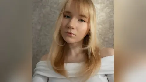 Blonde woman in a white off-the-shoulder top posing with a soft, serene expression against a textured wall.