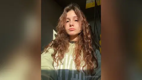 Young woman with long wavy hair wearing a sage green sweatshirt in warm sunlight.