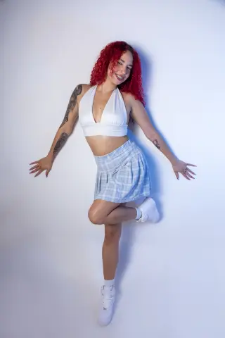 Smiling woman with curly red hair in a white crop top and blue plaid skirt posing against a white background.