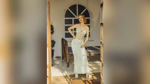 Elegant woman in a white lace maxi dress posing in a sunlit, minimalist modern room.
