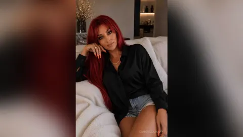 DonatellaRossy — DonatellaRossy with long red hair sits on a white sofa wearing a black button-down shirt and denim shorts.
