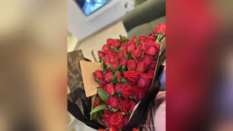 Large bouquet of fresh red roses wrapped in decorative paper held in a cozy indoor setting.