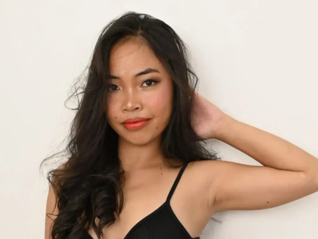Smiling young woman with long dark hair wearing a black camisole against a minimalist white background.