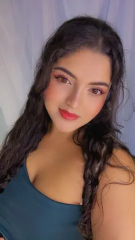 Young woman with long wavy hair wearing a dark teal tank top in a soft, bright indoor setting.