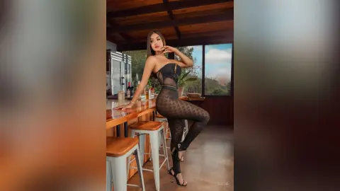 Glamorous woman in a black sheer bodysuit posing at a wooden bar in a modern luxury villa.