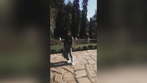 Stylish woman in a black leather jacket and jeans standing on a stone path in a sunny forest garden.