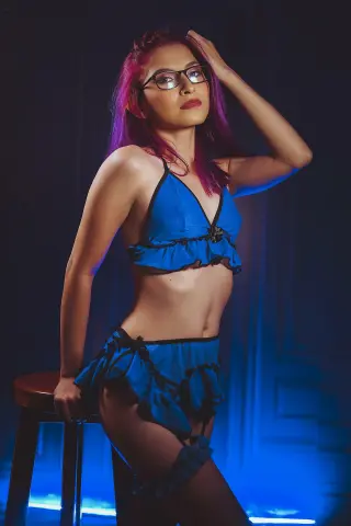 Young woman with pink hair in blue ruffled lingerie posing in moody blue studio lighting.