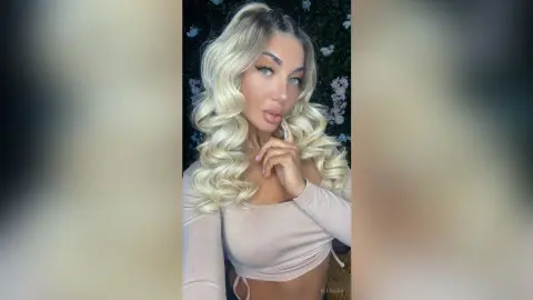 Blonde woman in a beige long-sleeve top posing with a glamorous look against a floral background.