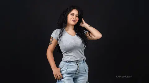 KaiaSinclair poses with long dark hair, wearing a tight grey t-shirt and light blue jeans against a black background.