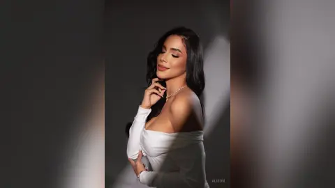 Alison poses with her eyes closed, wearing a white off-the-shoulder top and a gold necklace against a dark background.