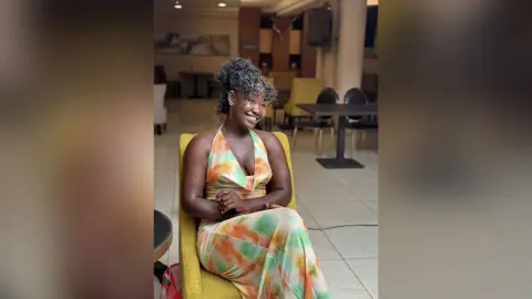 Smiling Black woman in a colorful floral maxi dress sitting in a modern lounge setting.