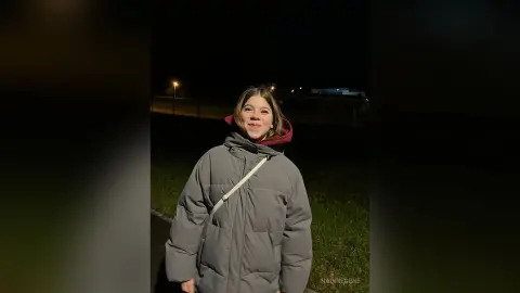 Smiling young woman in a grey puffer jacket standing outdoors at night under dim streetlights.
