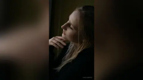 Melancholic blonde woman in a black outfit looking out a window in soft, moody indoor lighting.