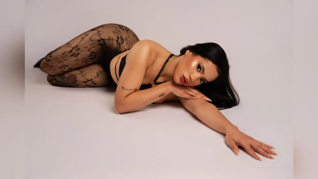 GiaDuval poses in a white bathtub wearing black fishnet lingerie against a red background.