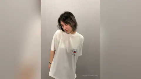 Young woman with short dark hair wearing a white graphic t-shirt in a minimalist indoor setting.