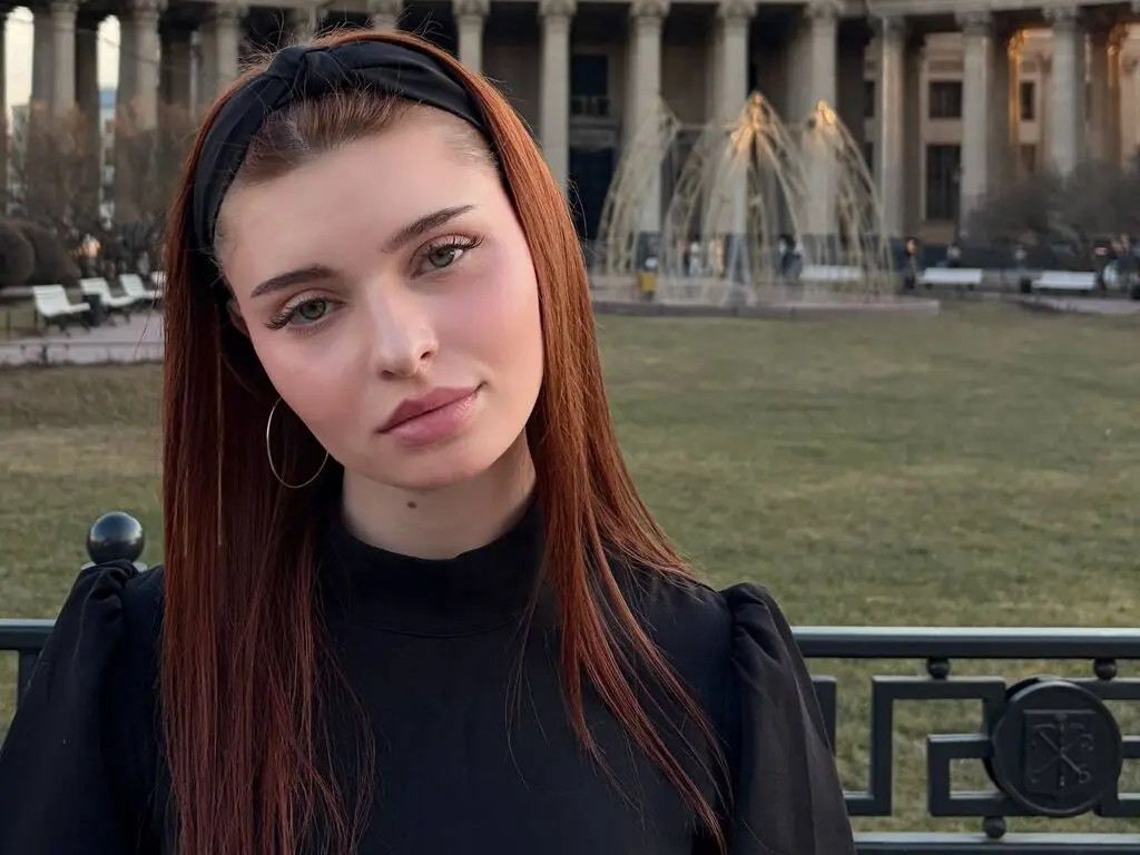 Redheaded woman in a black turtleneck and headband sitting on a park bench near a fountain at sunset.